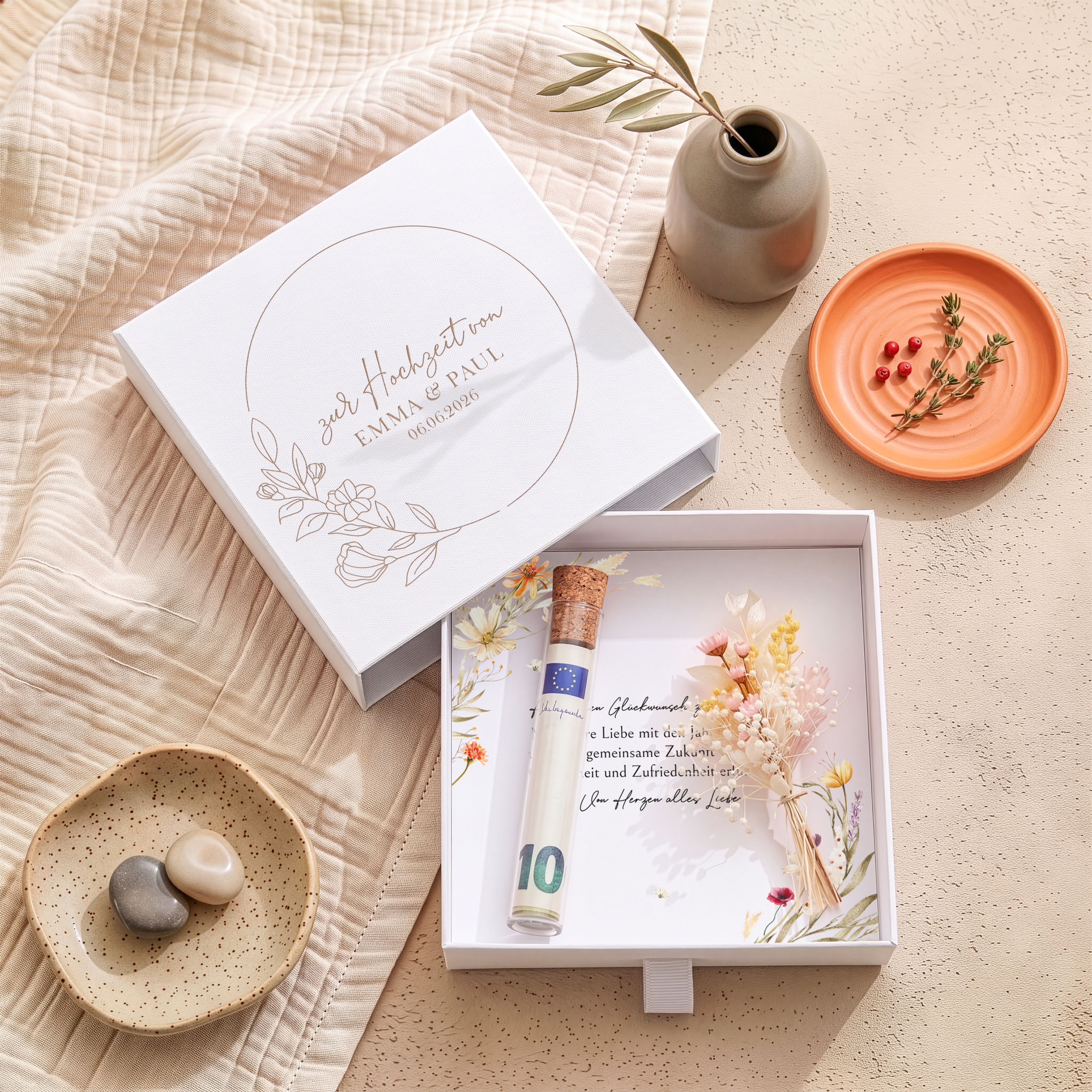 Weiße personalisierte Hochzeits-Geschenkbox aus Karton. Der Deckel zeigt eine goldene Blütenkranz-Gravur mit Namen und Datum. Innen liegen eine floral bedruckte Karte, ein Röhrchen und Trockenblumen.