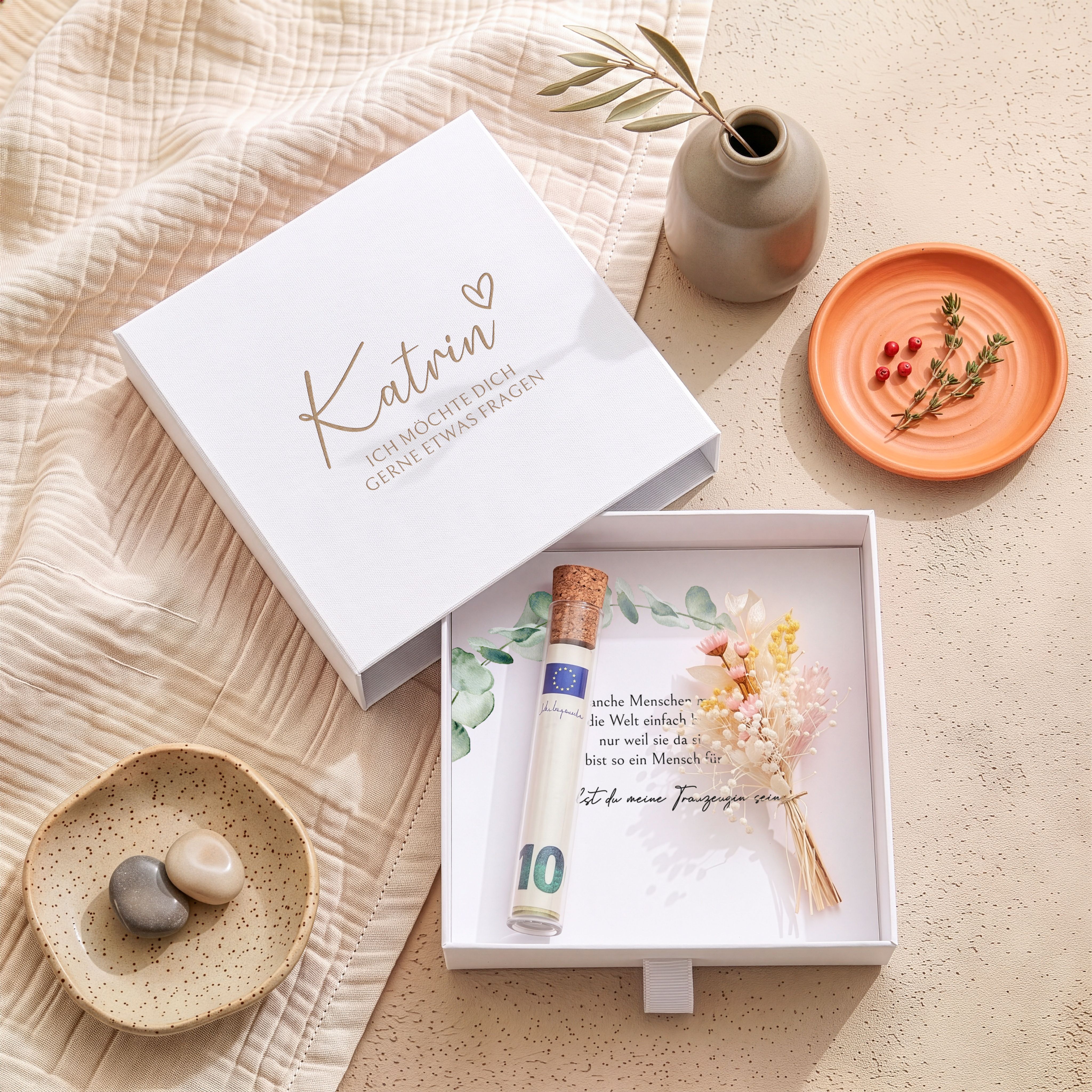 Weiße Geschenkbox aus Karton zur Trauzeugenfrage. Der Deckel ist mit goldener Namensgravur und Herz verziert. Innen liegen ein Glasröhrchen, eine bedruckte Einlegekarte und ein rosa-beiges Trockenblumensträußchen.