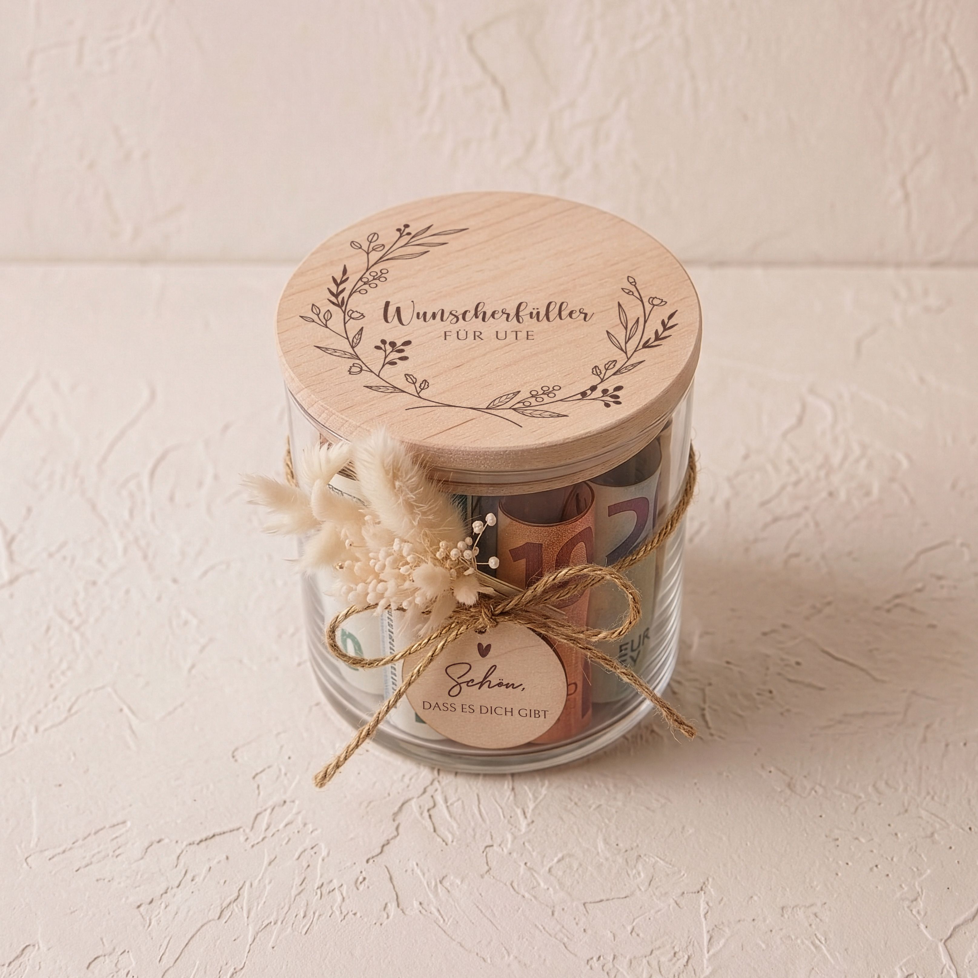 Vorratsglas mit graviertem Buchenholzdeckel und Blütenkranz-Motiv. Eine Kordel hält einen Holzanhänger und ein beiges Trockenblumensträußchen.