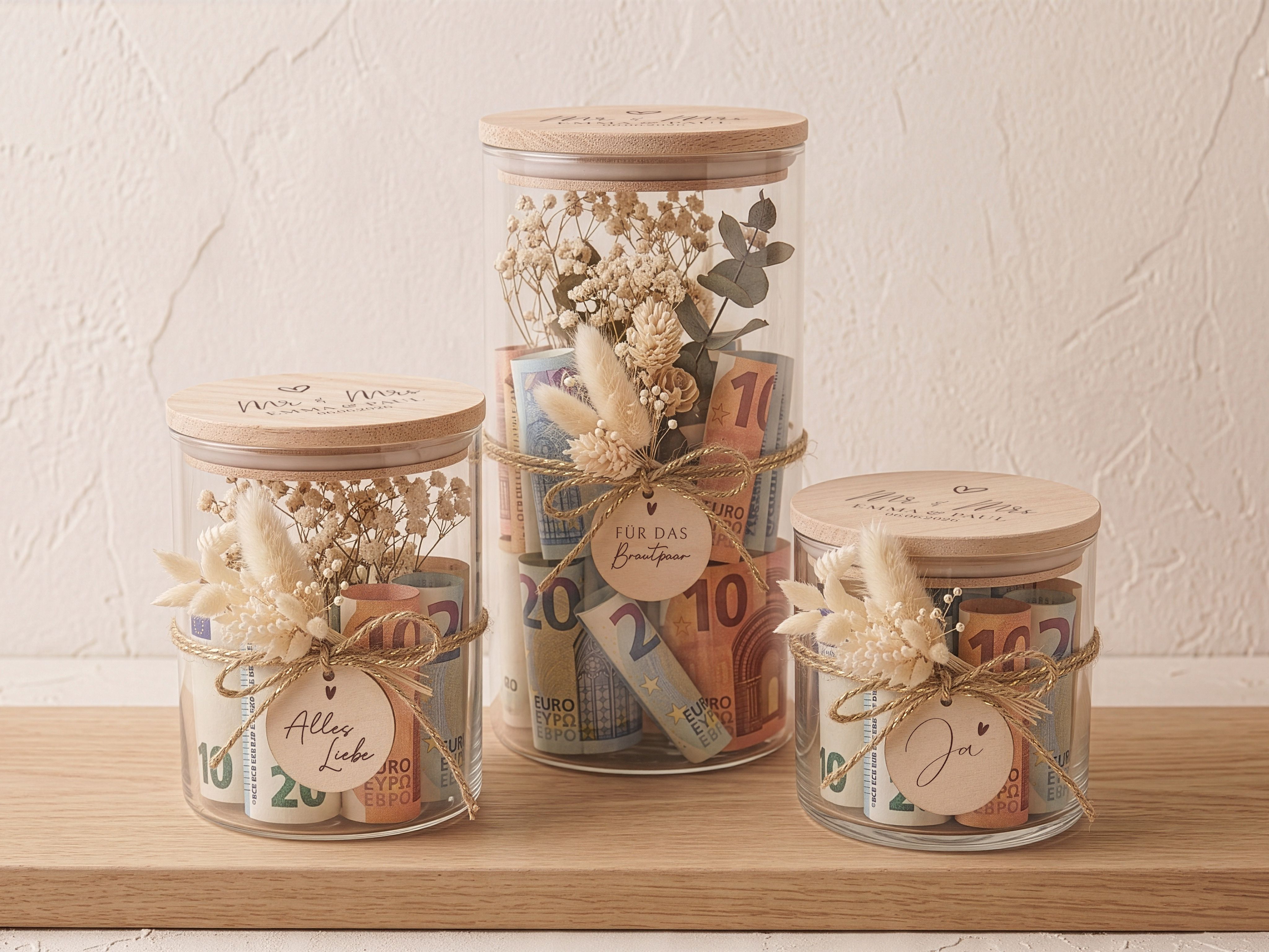 Personalisierte Vorratsgläser aus Glas mit graviertem Buchenholzdeckel für Hochzeitsgeschenke. Das Design zeigt einen 'Mr & Mrs' Schriftzug. Sichtbar sind beige Trockenblumensträußchen und runde Holzanhänger.