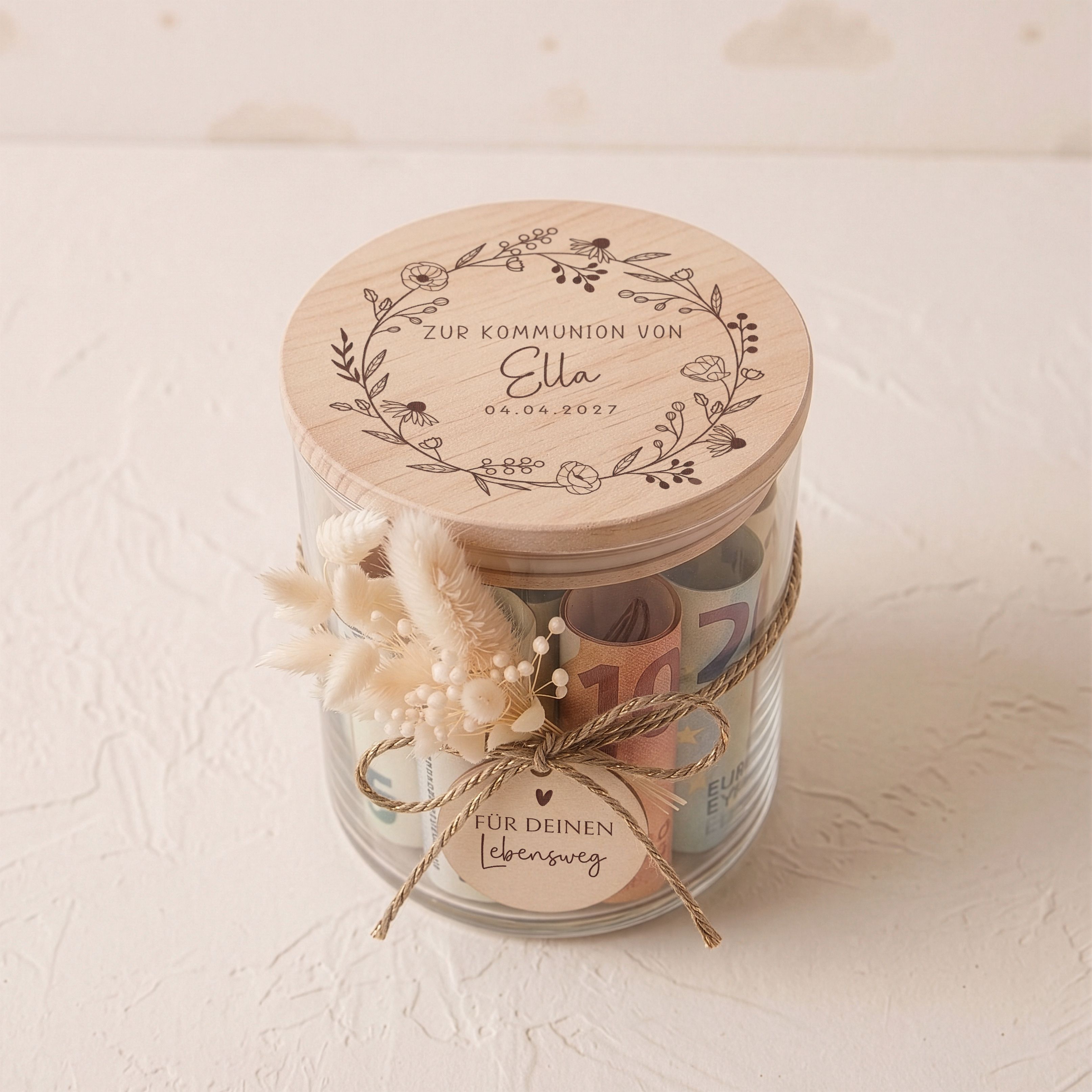 Personalisiertes Vorratsglas zur Kommunion mit graviertem Holzdeckel und Blütenkranz. Beige Trockenblumen und ein Anhänger 'Für deinen Lebensweg' schmücken das Glas.