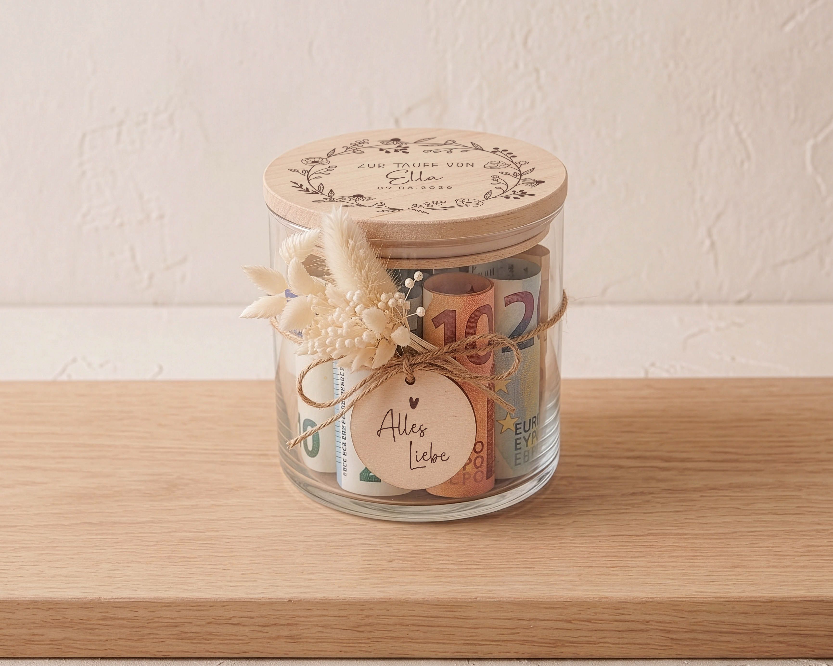 Personalisiertes Glasgefäß zur Taufe mit graviertem Buchenholzdeckel und Blütenkranz. Ein Holzanhänger 'Alles Liebe' und beige Trockenblumen sind befestigt.