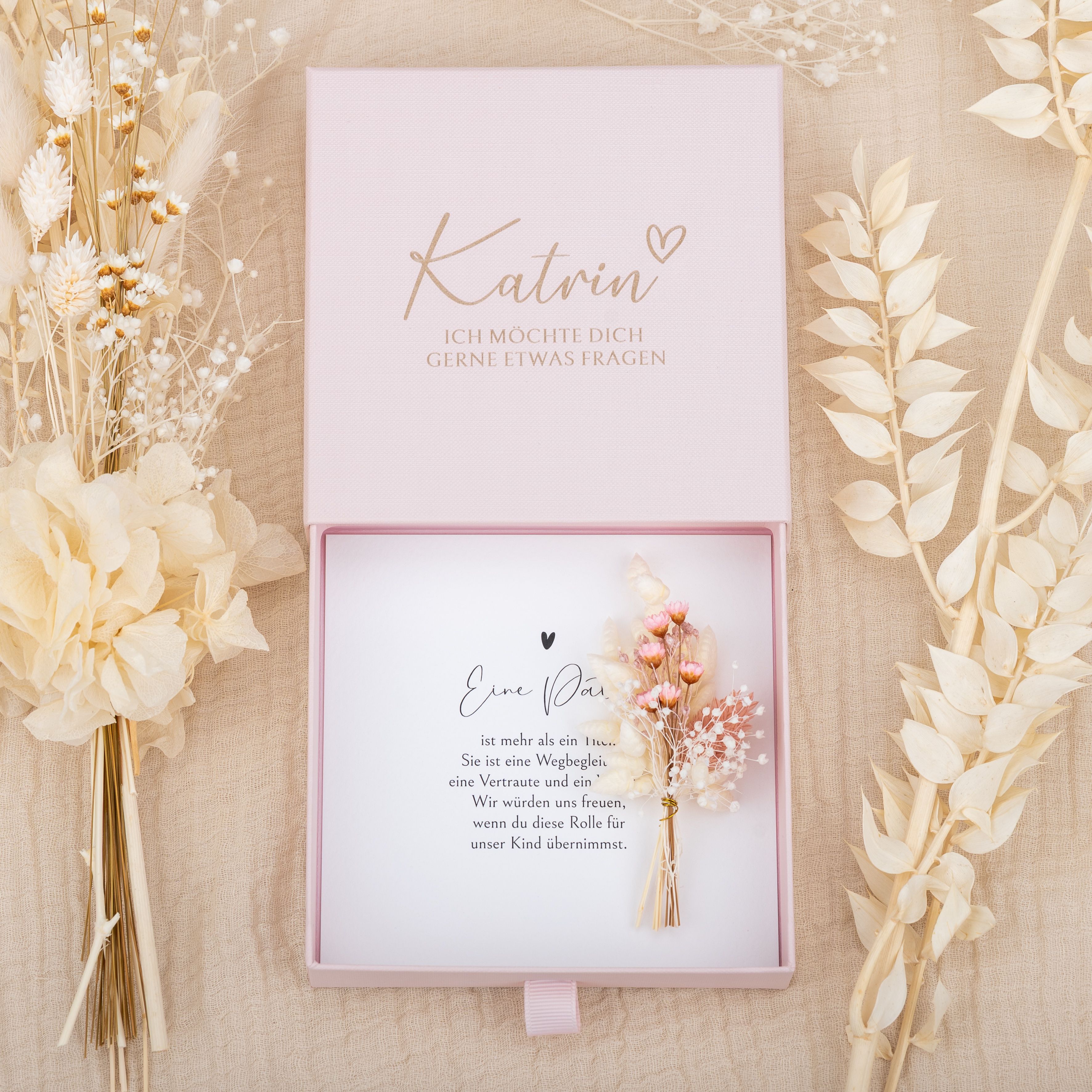 Personalisierte Geschenkbox aus rosa Karton zur Patenfrage. Deckel mit graviertem Namen Katrin und Herz. Innen Trockenblumen.